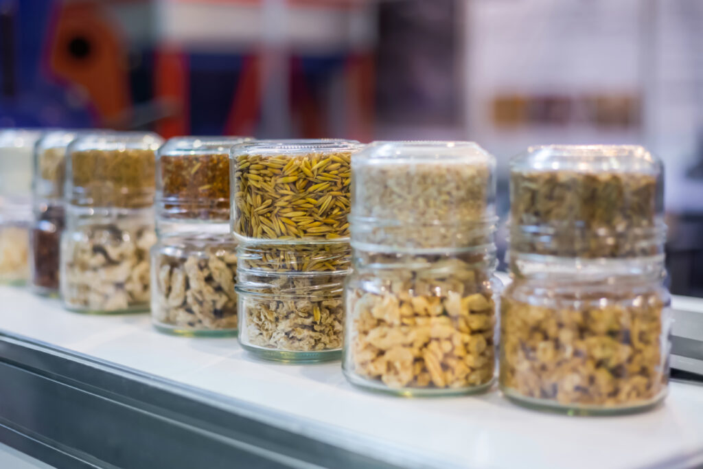 Different kinds of pelleted compound feed, fodder in glass jars: before and after extrusion - food for domestic animals at agricultural exhibition, trade show: close up. Agriculture, husbandry concept